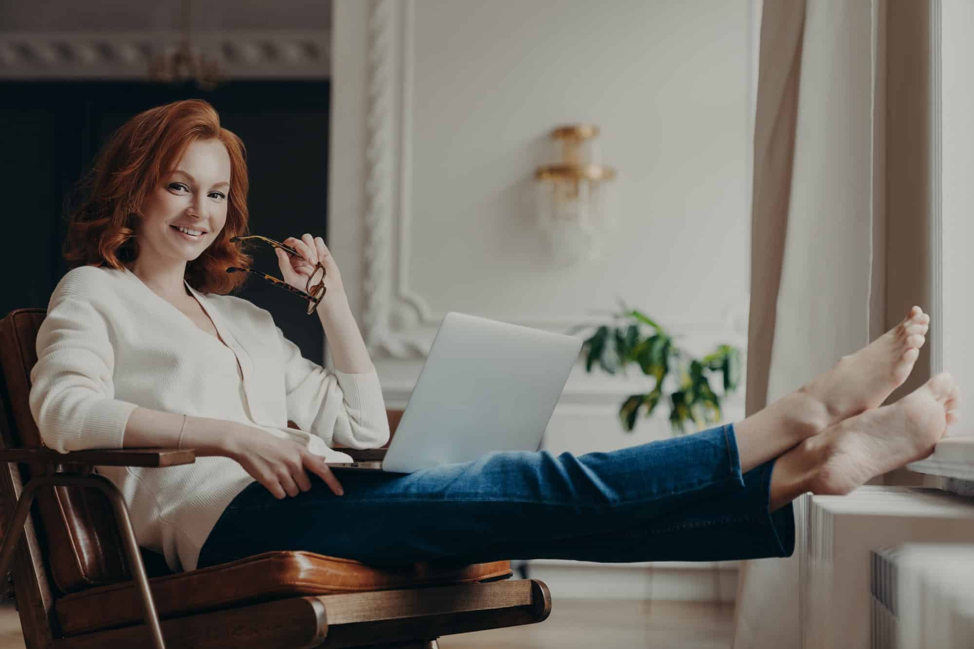 Cheerful successful ginger woman copywriter works from home, keeps laptop on knees, stretches legs